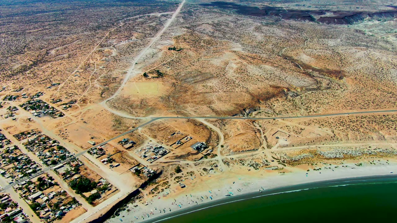 A wide aerial frame over San Juanico, Baja California Sur, showing green-blue water, a sandy beach with tiny visible people, clustered homes, dirt roads, and arid desert beyond.