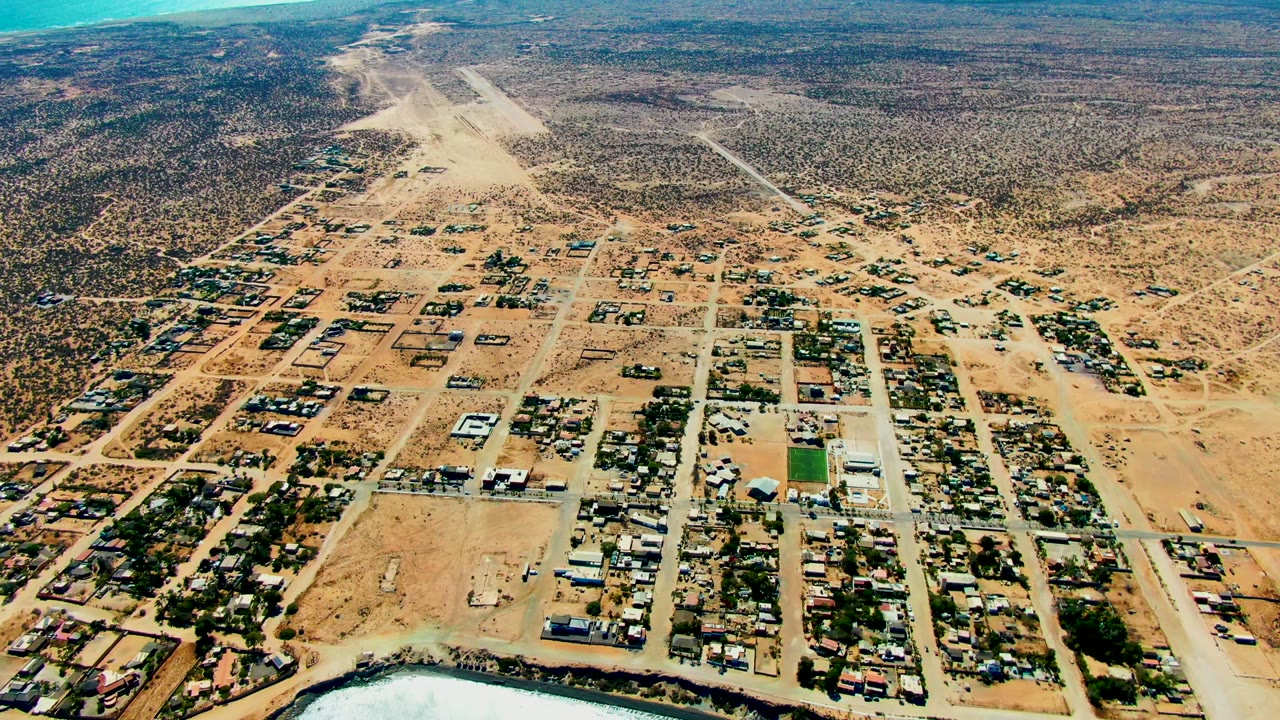 Wide aerial frame over San Juanico, Baja California Sur, showing grid roads, low buildings, sandy terrain, desert scrub, green water at the lower edge, and no visible people.