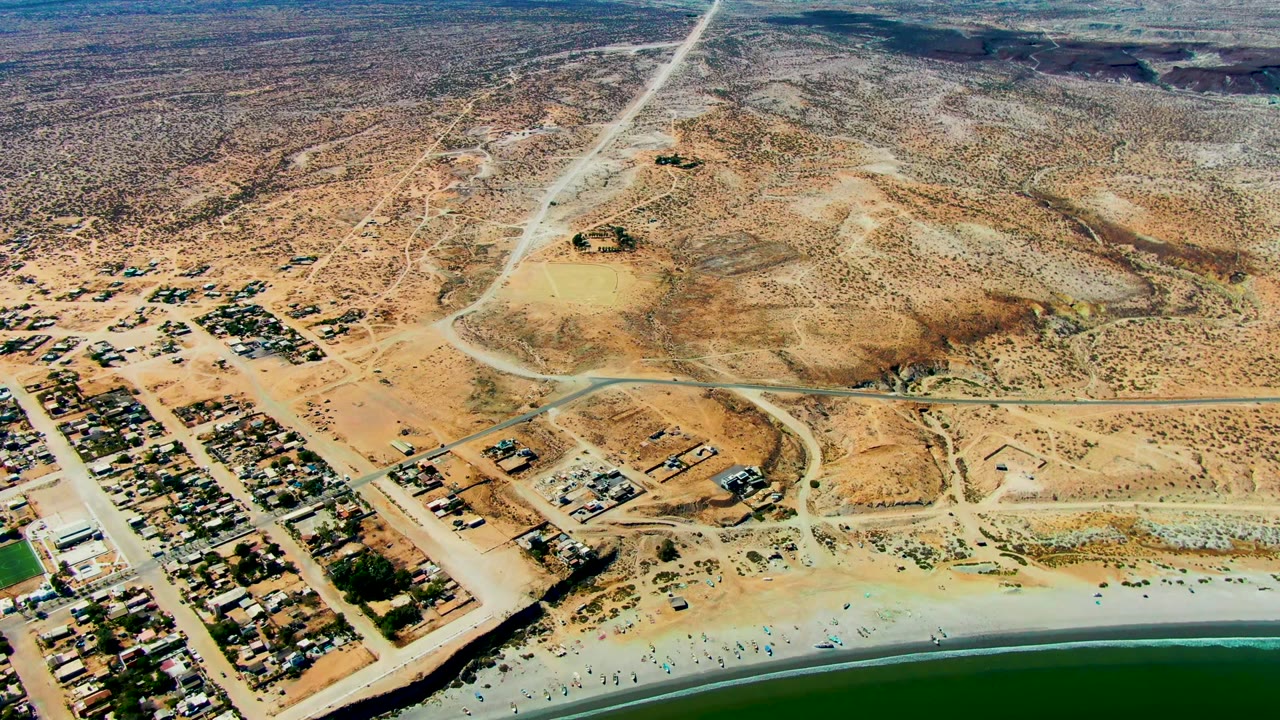High aerial frame of San Juanico showing a cluster of buildings near the shore, curving dirt roads through tan desert, low hills, green water, and no visible people.