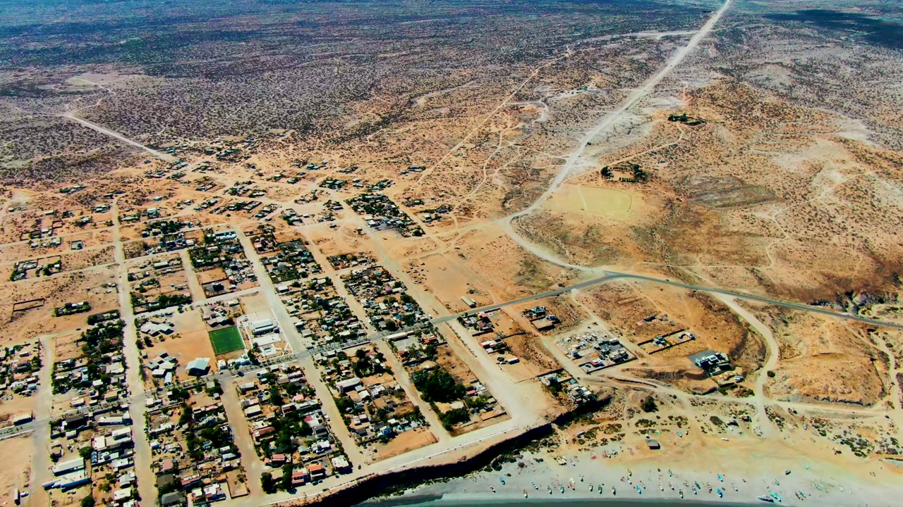High aerial view over San Juanico, Baja California Sur, showing sandy streets, small buildings, desert scrub, shoreline surf, and no visible people.