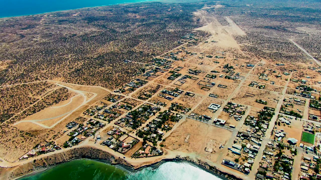 Oblique aerial view in San Juanico showing tan town blocks, a low scrub-covered rise, pale beach, turquoise water, and no visible people.