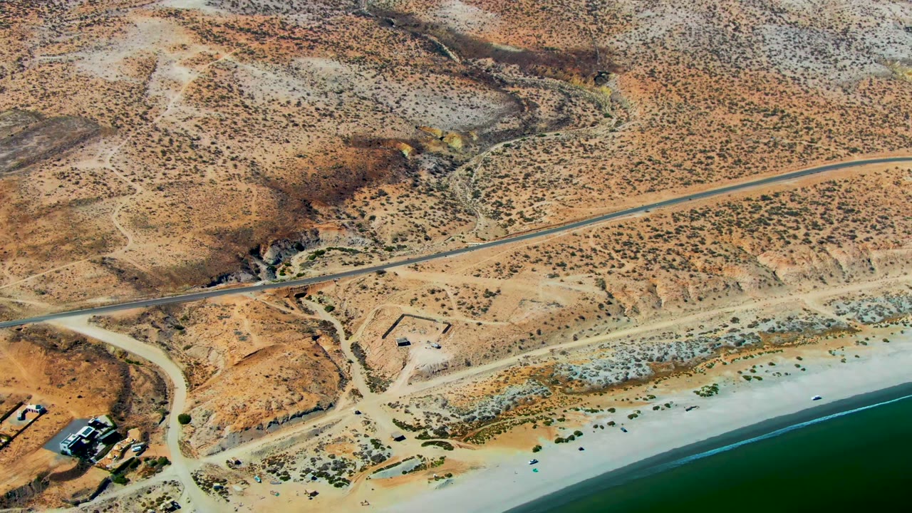 Aerial view over San Juanico, Baja California Sur, showing dry hills, a road above the beach, pale sand, green ocean water, and no visible people.