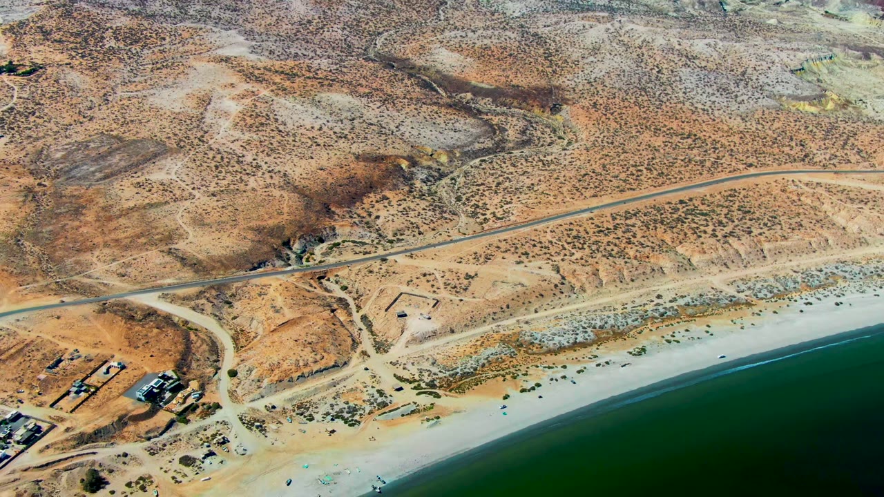 Aerial coastline with arid terrain, beach, road, and small building cluster