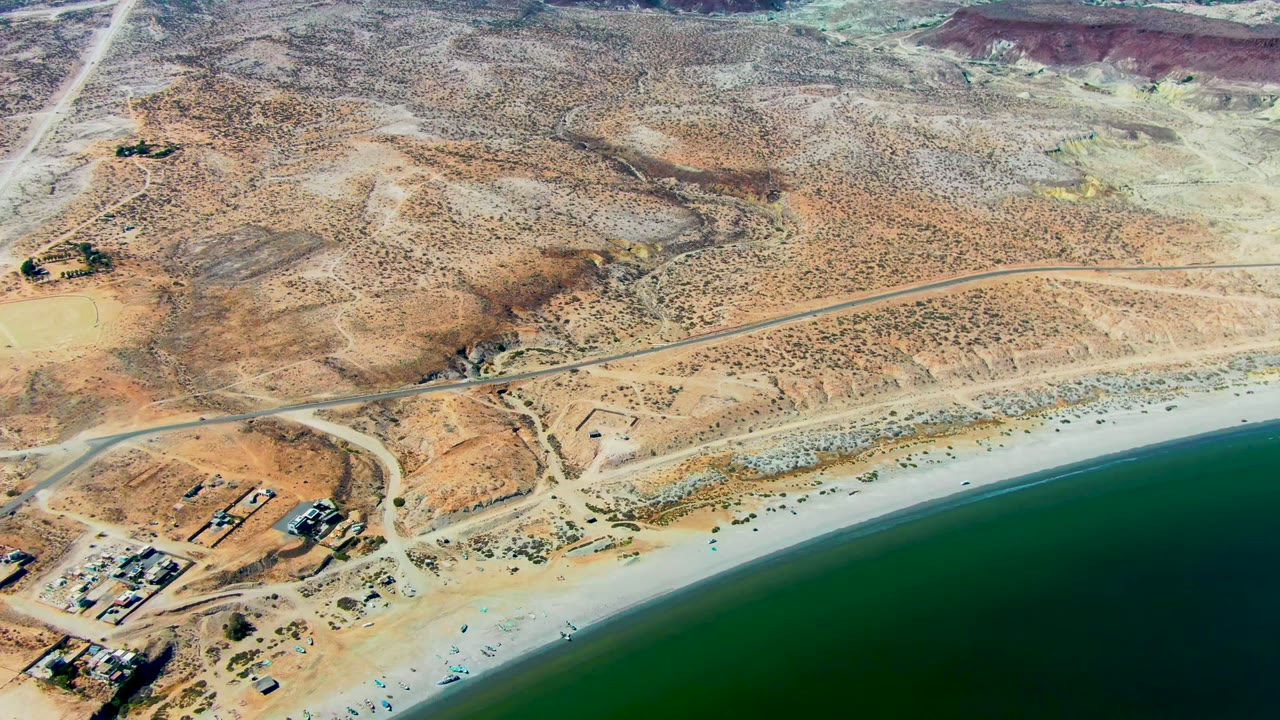 Drone view over San Juanico with a curving pale beach, green water, dry hills, shoreline roads, scattered structures, and no visible people.