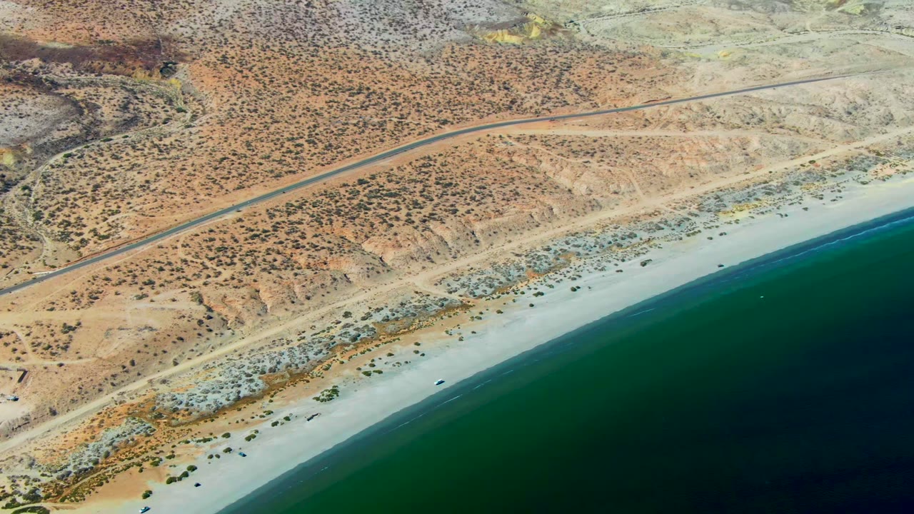 High aerial view over San Juanico, Baja California Sur, with a paved road above a pale beach, dark green ocean along the lower right, sparse desert scrub, and no visible people.