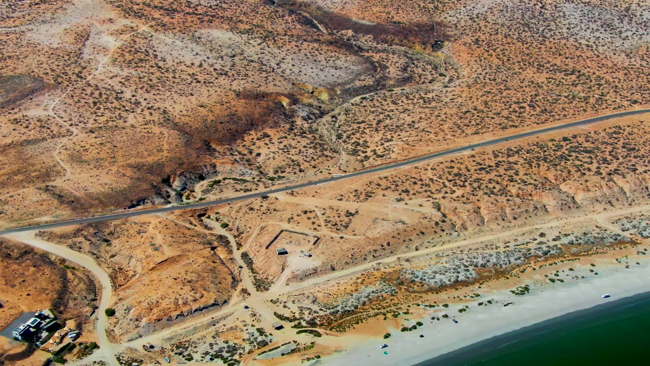 Drone view over San Juanico with rust-colored hills, a paved road, branching dirt tracks, a sandy beach, green ocean water, small shoreline structures, and no visible people.