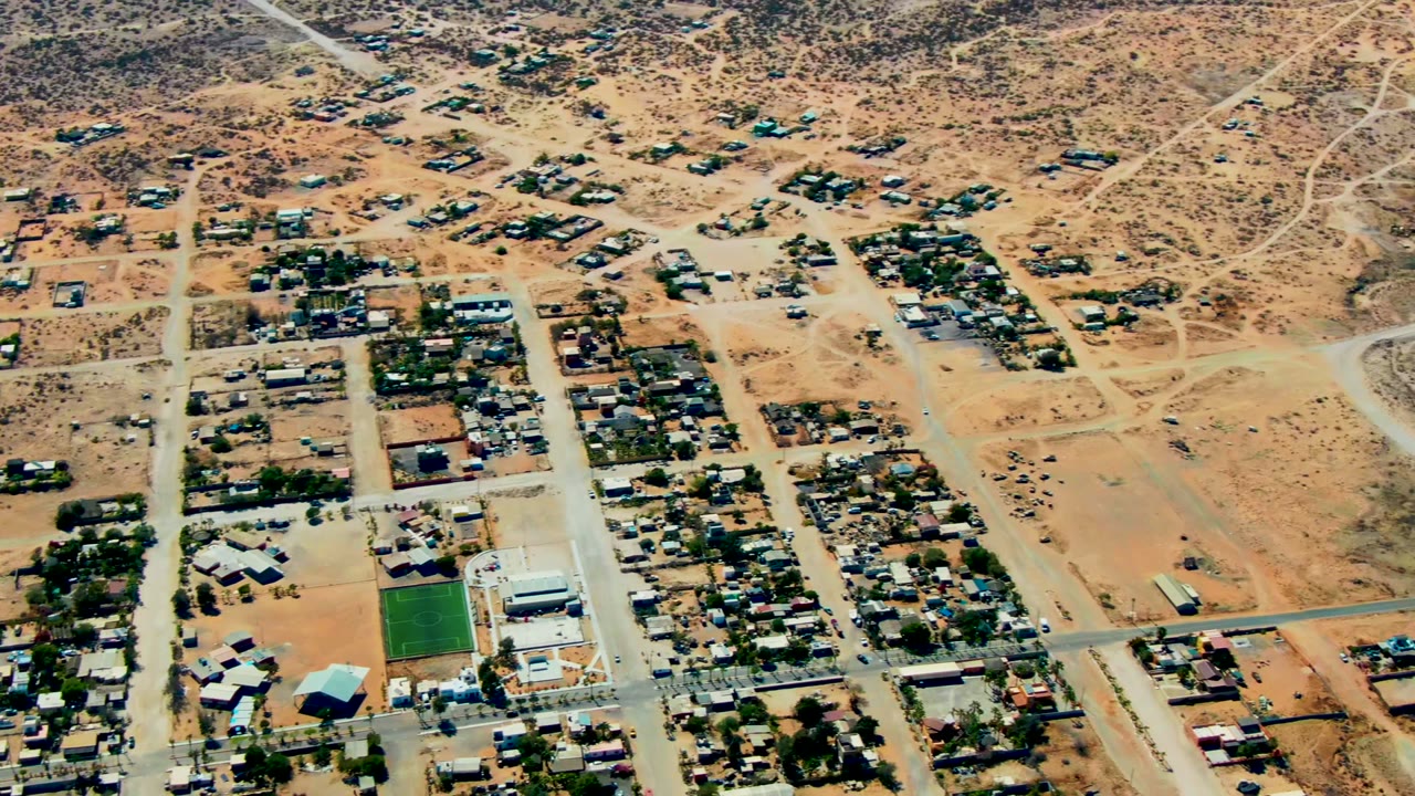 Wide aerial view in San Juanico, Baja California Sur showing clustered homes, rectilinear roads, dry lots, desert hills beyond, and no visible people.