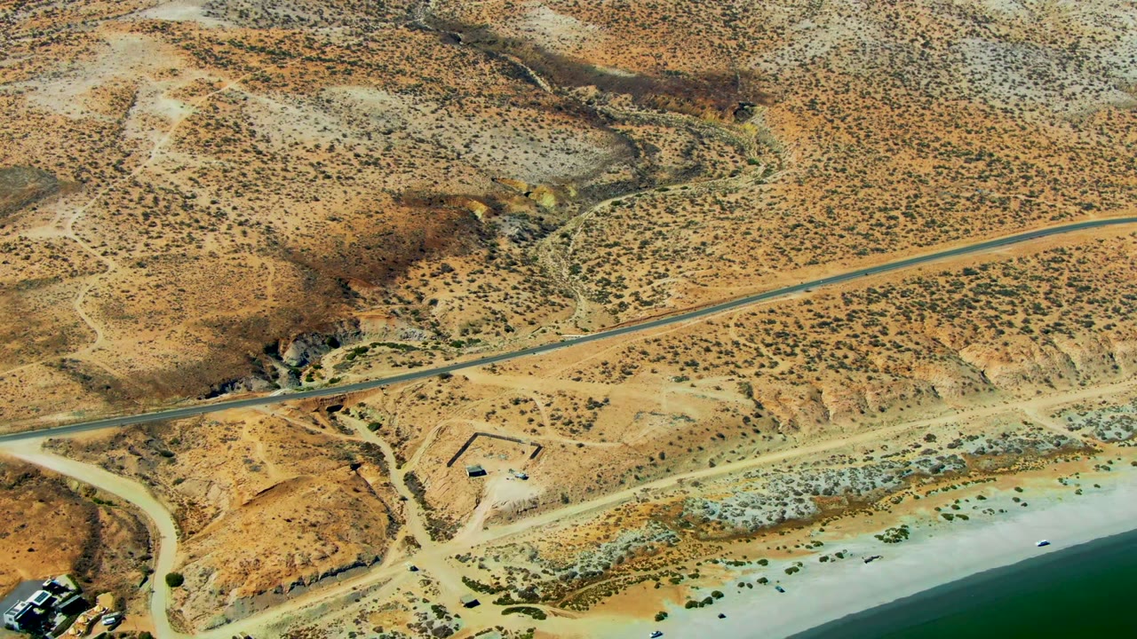 High aerial view over San Juanico showing tan desert hills, a road, pale beach, green ocean water, scattered tracks, and no visible people.