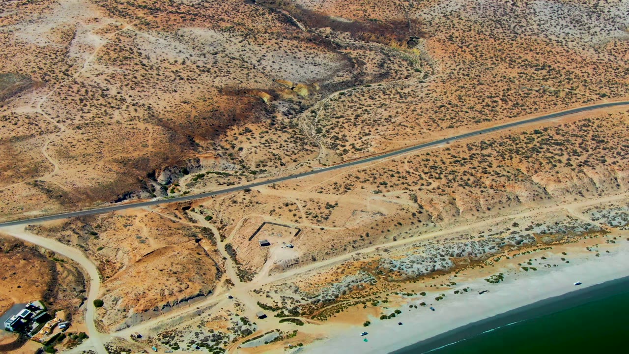 High overhead view in San Juanico where a paved road crosses dry terrain above a pale beach and green shoreline, with no visible people.
