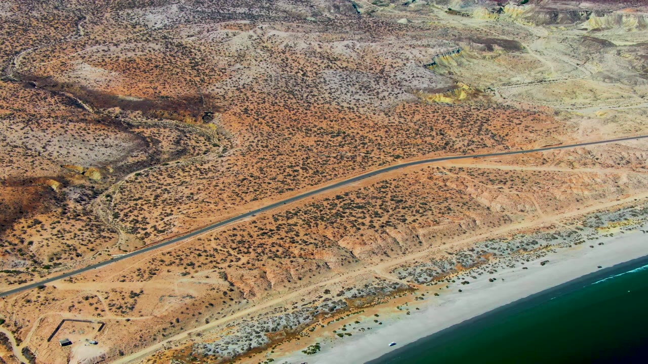 High aerial view in San Juanico, Baja California Sur, with a paved road running across scrubby desert above a pale sandy beach and blue-green ocean, no visible people.