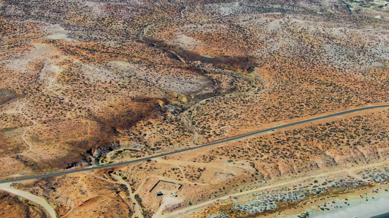 Aerial frame in San Juanico with the road crossing near a dry wash, a small rectangular site below it, and the beach and ocean along the lower right, no visible people.