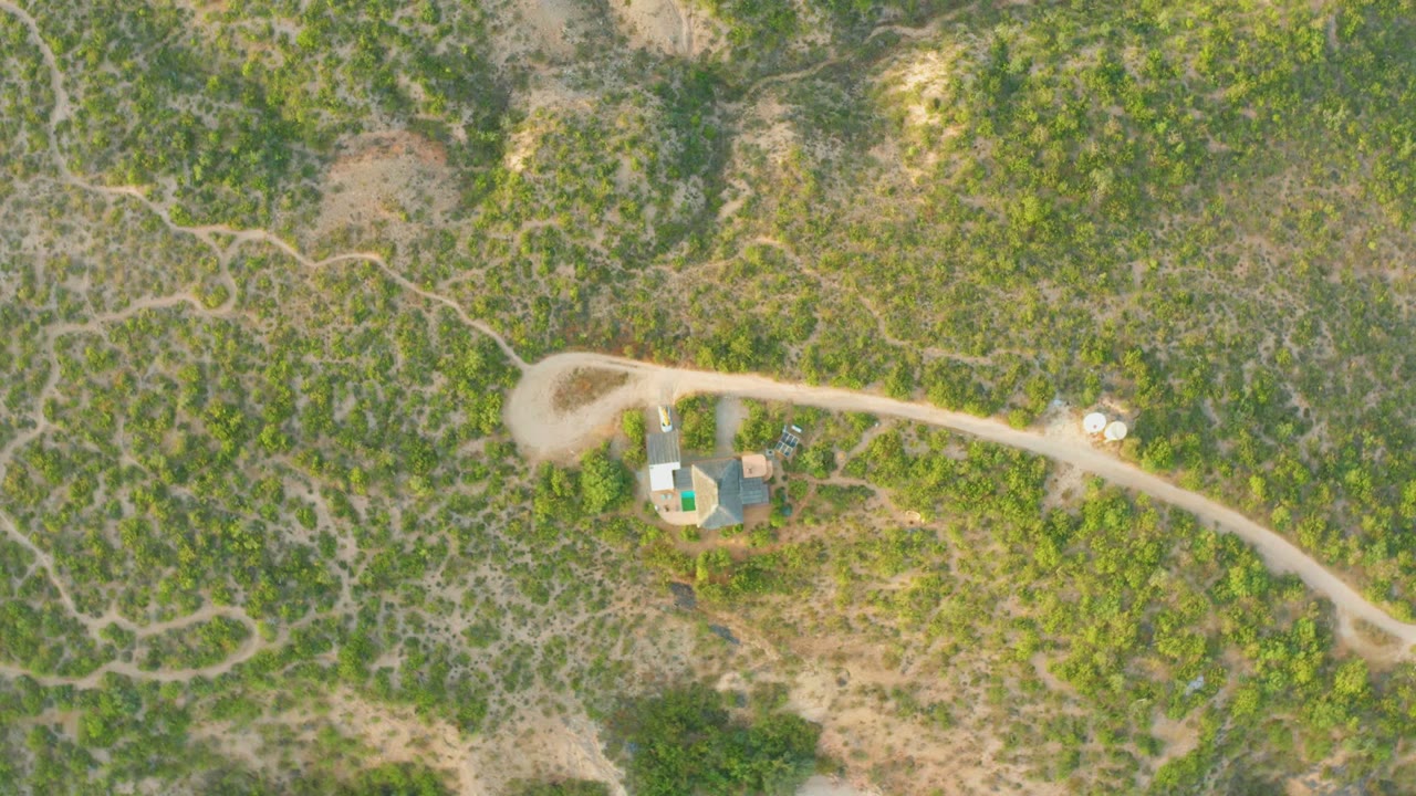 Aerial view of a vehicle on a pale dirt road beside small structures and scrub hills.