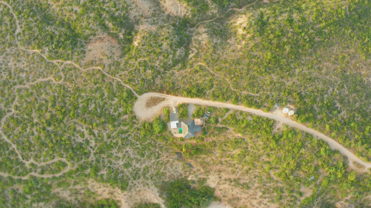 High overhead view in Cabo Pulmo with the vehicle continuing along the road, low green vegetation across the hills, dry tan ground, and no visible people.
