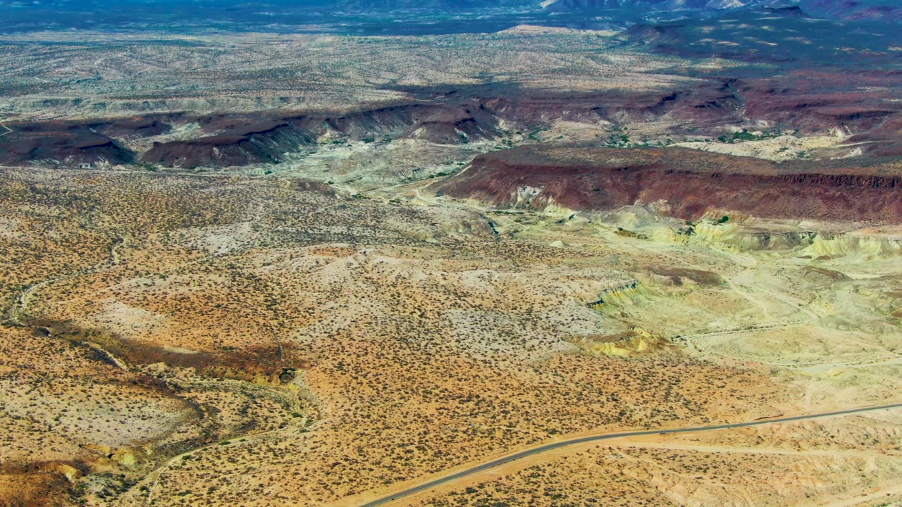 Wide aerial view over San Juanico desert terrain with a road crossing the lower frame, scrub-covered flats, pale eroded hills, dark ridges, and no visible people.