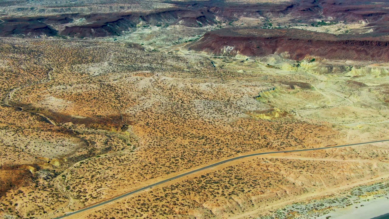 Aerial view over San Juanico desert with rocky washes, a road running across the lower frame, the first strip of beach and green-blue water entering at the right, and no visible people.