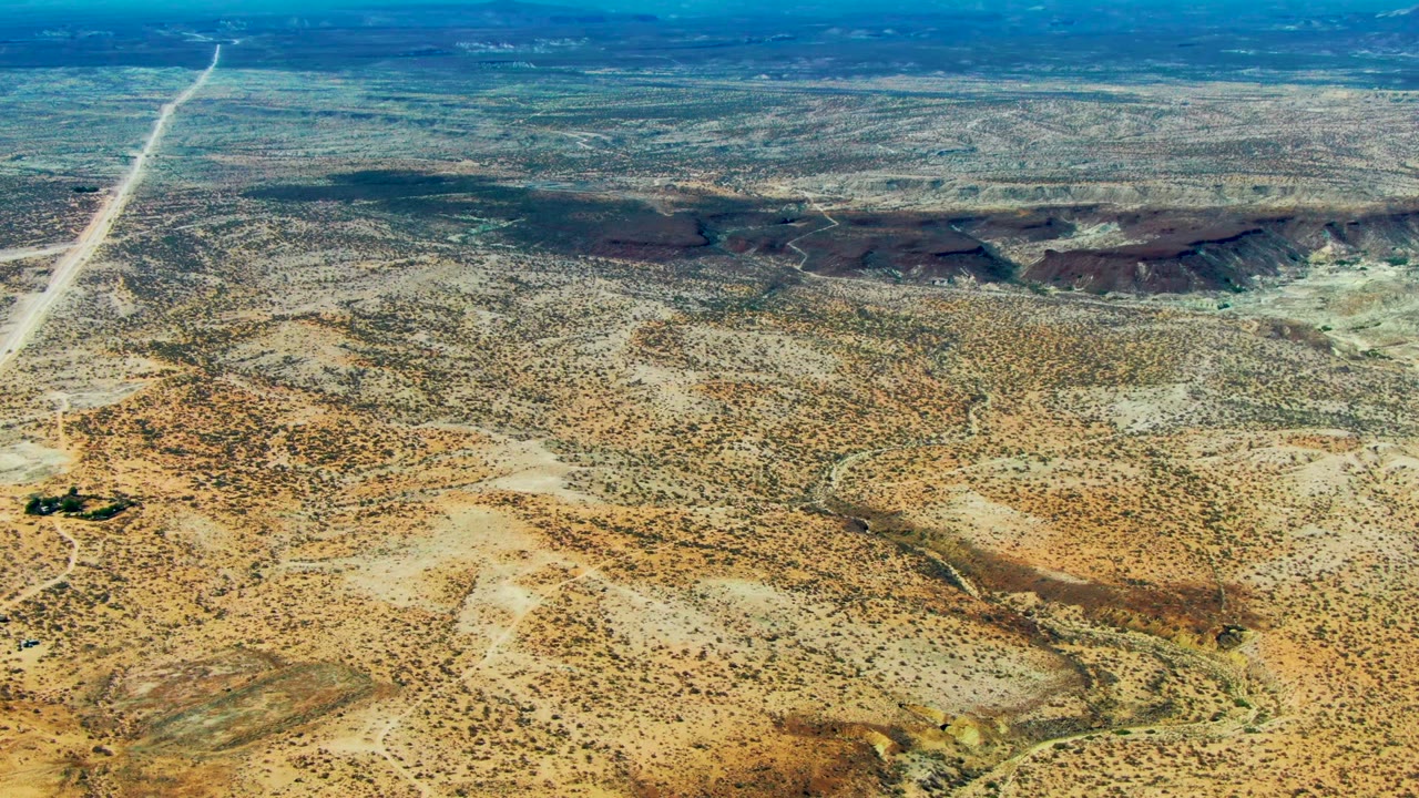 High aerial view over San Juanico desert terrain with a straight pale road cutting through scrub and low dark hills in the distance, no visible people.