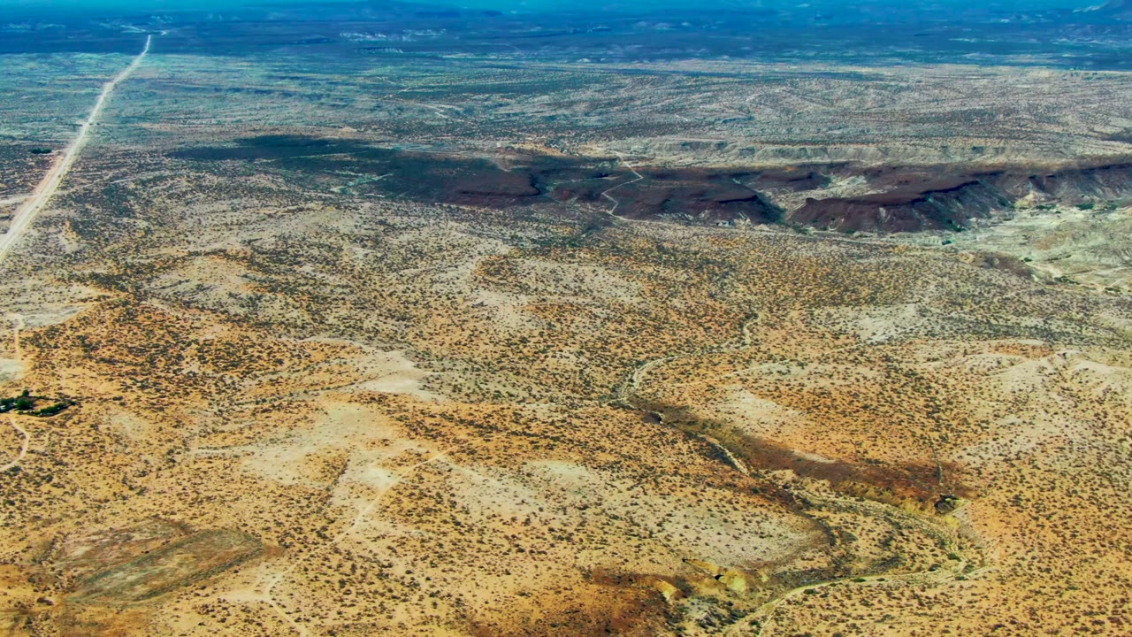 Wide aerial frame over San Juanico desert with a bright road running toward low dark ridges and mottled scrub across the foreground, no visible people.