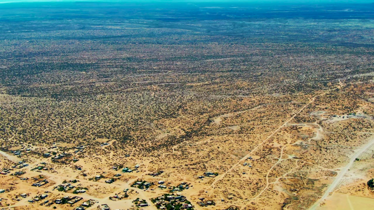 Wide aerial view over San Juanico, Baja California Sur desert with pale dirt roads, sparse scrub, scattered structures at lower left, distant ridges, and no visible people.