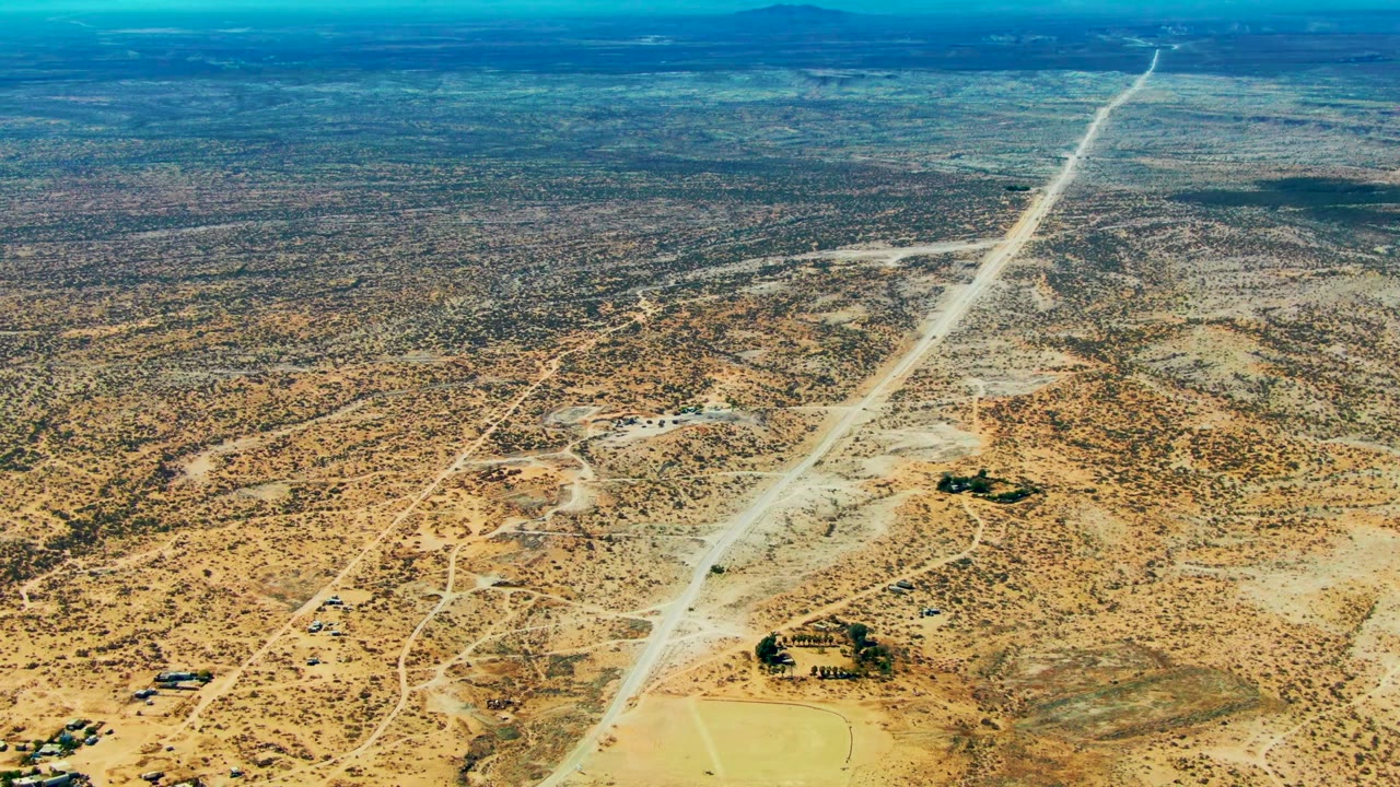 High aerial view over San Juanico, Baja California Sur, with a pale dirt road running through sandy scrubland toward distant low hills and no visible people.
