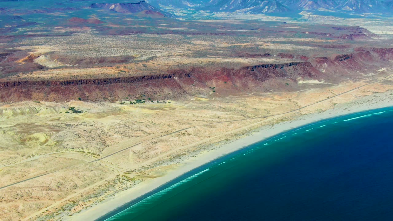 Aerial frame over San Juanico, Baja California Sur showing blue ocean along a sandy shoreline, pale desert flats, red rock ridges, distant mountains, and no visible people.