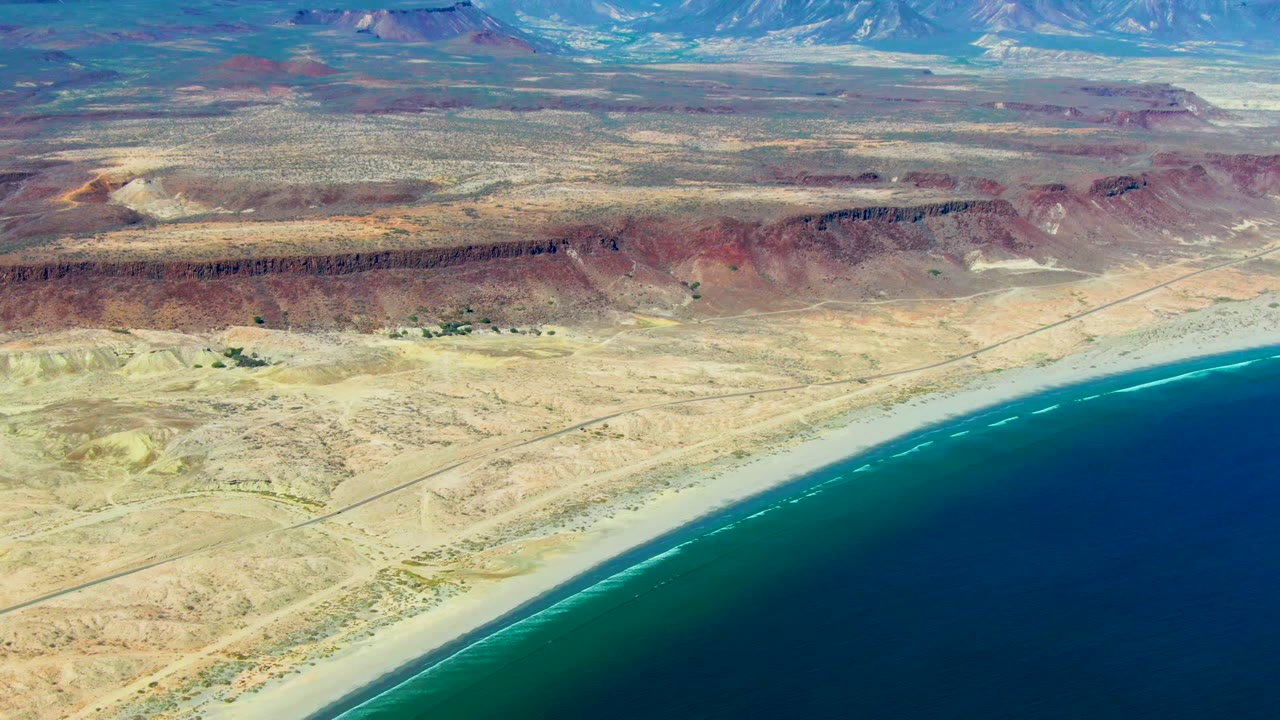 Aerial coast view with turquoise surf, sandy desert, red ridge, road lines, and mountains.