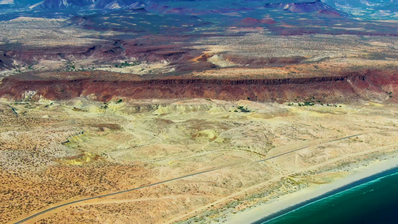 Aerial frame over San Juanico, Baja California Sur showing the ocean pushed to the lower right edge, a road crossing sandy desert, red rock formations, and no visible people.