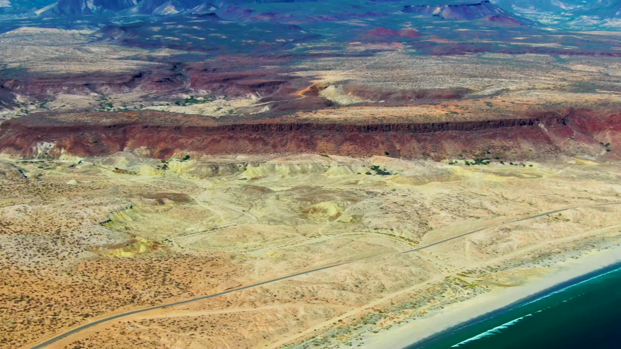 Aerial frame over San Juanico, Baja California Sur, showing turquoise ocean, a pale beach, a parallel road, tan desert, red cliffs, distant mountains, and no visible people.