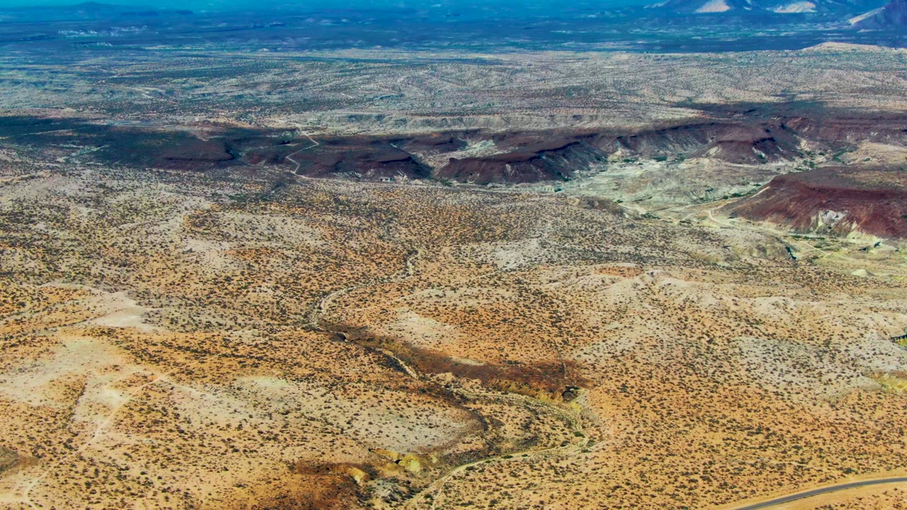 Aerial view over scrubby desert with cliffs and distant mountains.