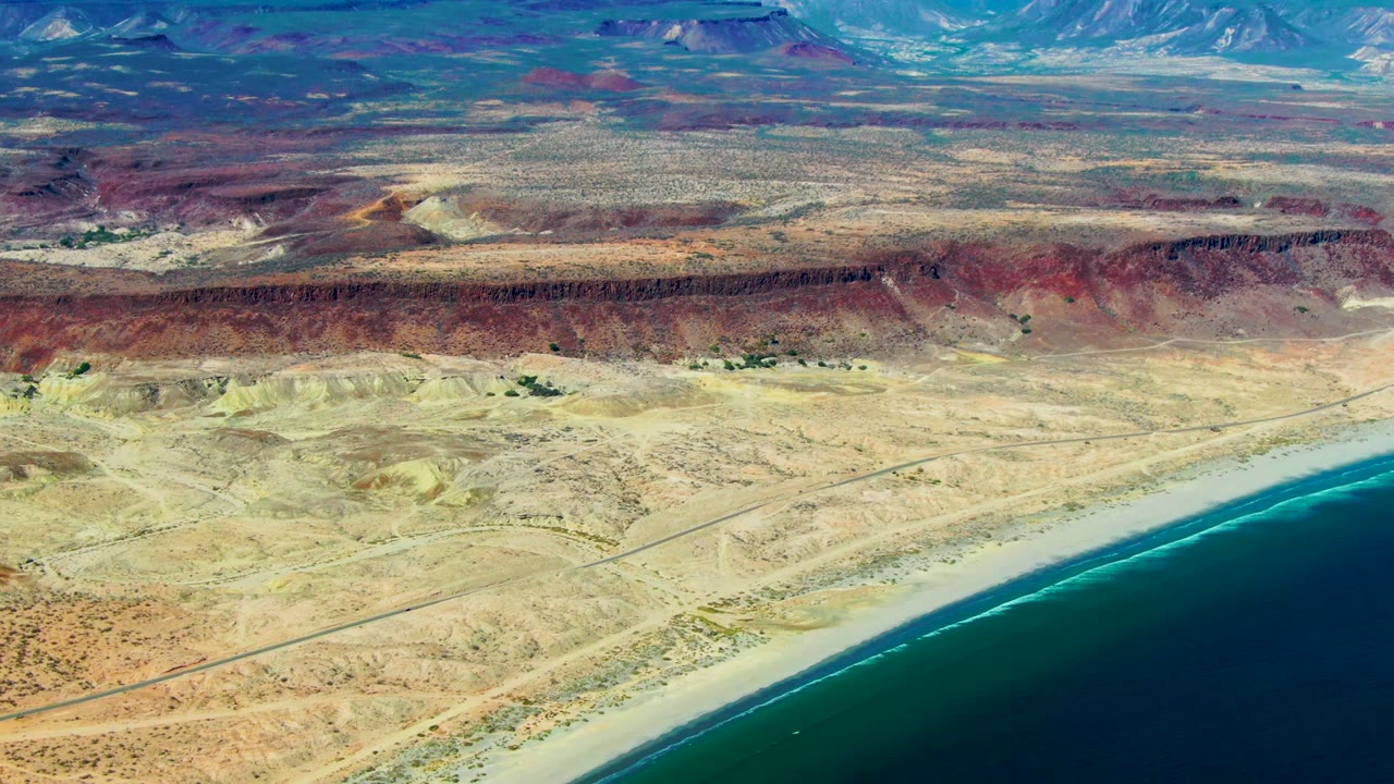 Opening aerial frame over San Juanico, Baja California Sur, with dark ocean at lower right, a sandy beach, tan desert flats, reddish bluffs, distant mountains, and no visible people.