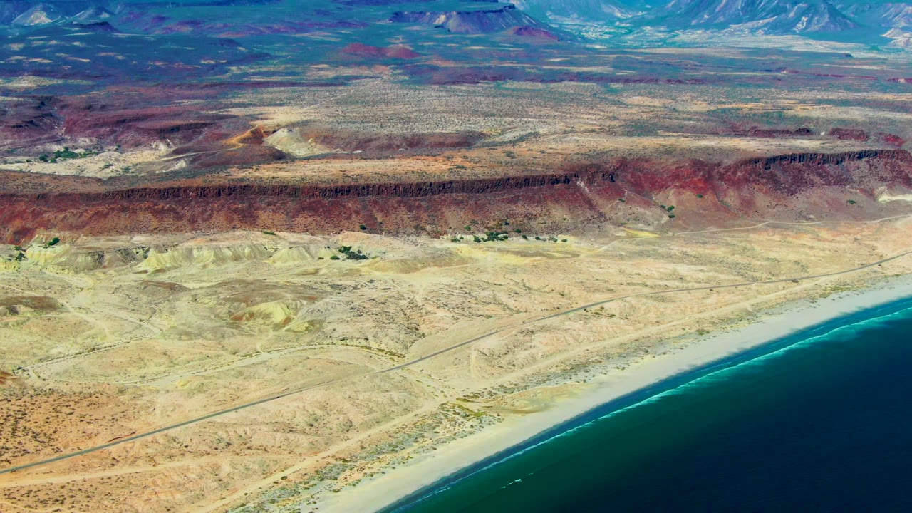 Wide aerial coastline with desert flats, red bluff band, beach, and ocean.