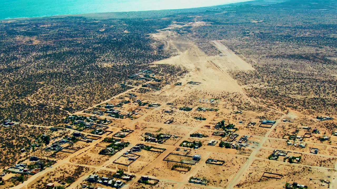 High aerial frame over San Juanico, Baja California Sur, showing scattered houses, pale dirt roads, scrubby desert, blue ocean on the left horizon, distant hills, and no visible people.