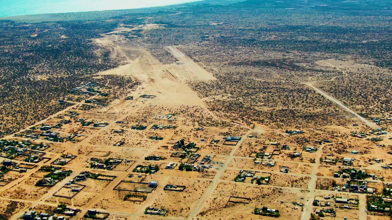 Drone view of sandy roads, small buildings, desert scrub, and blue ocean horizon.