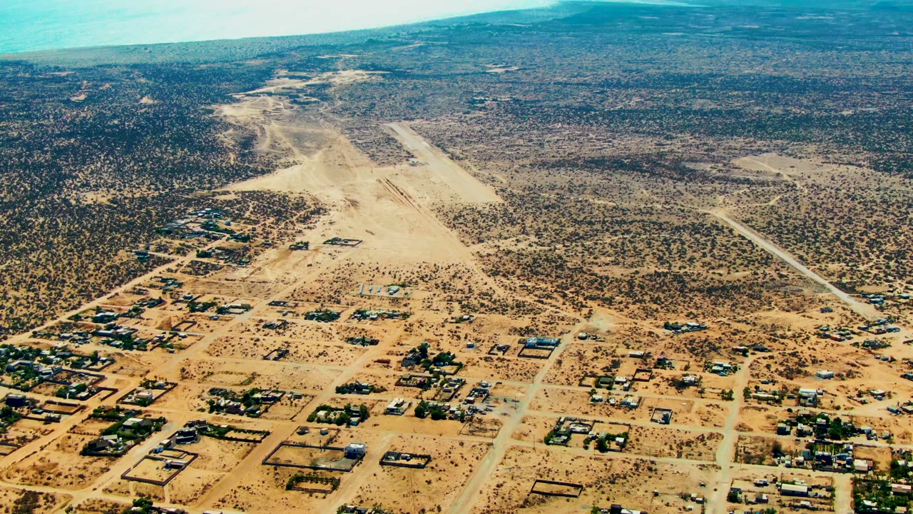 High drone frame over San Juanico, Baja California Sur, showing a long straight dirt road at right, clustered houses below, broad scrubland, blue ocean across the upper left, and no visible people.