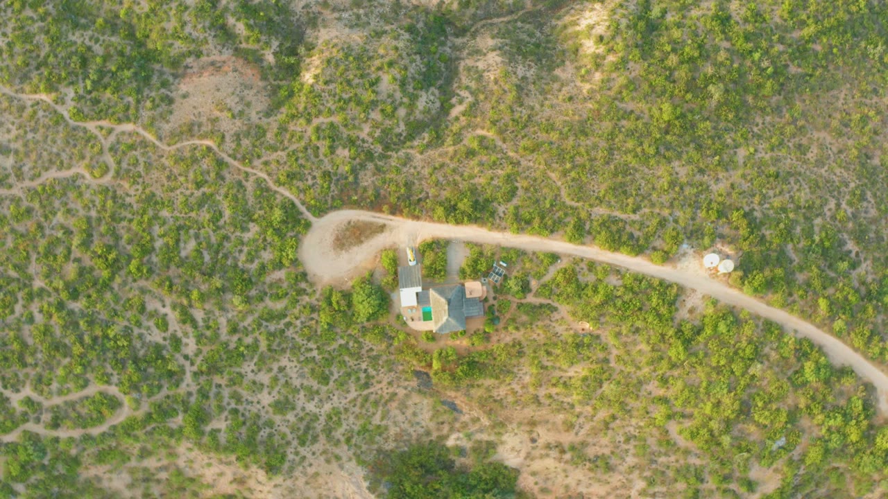 Aerial frame over Cabo Pulmo, Baja California Sur, showing a pale dirt road, small buildings, mixed green and brown hillside vegetation, and no visible people.