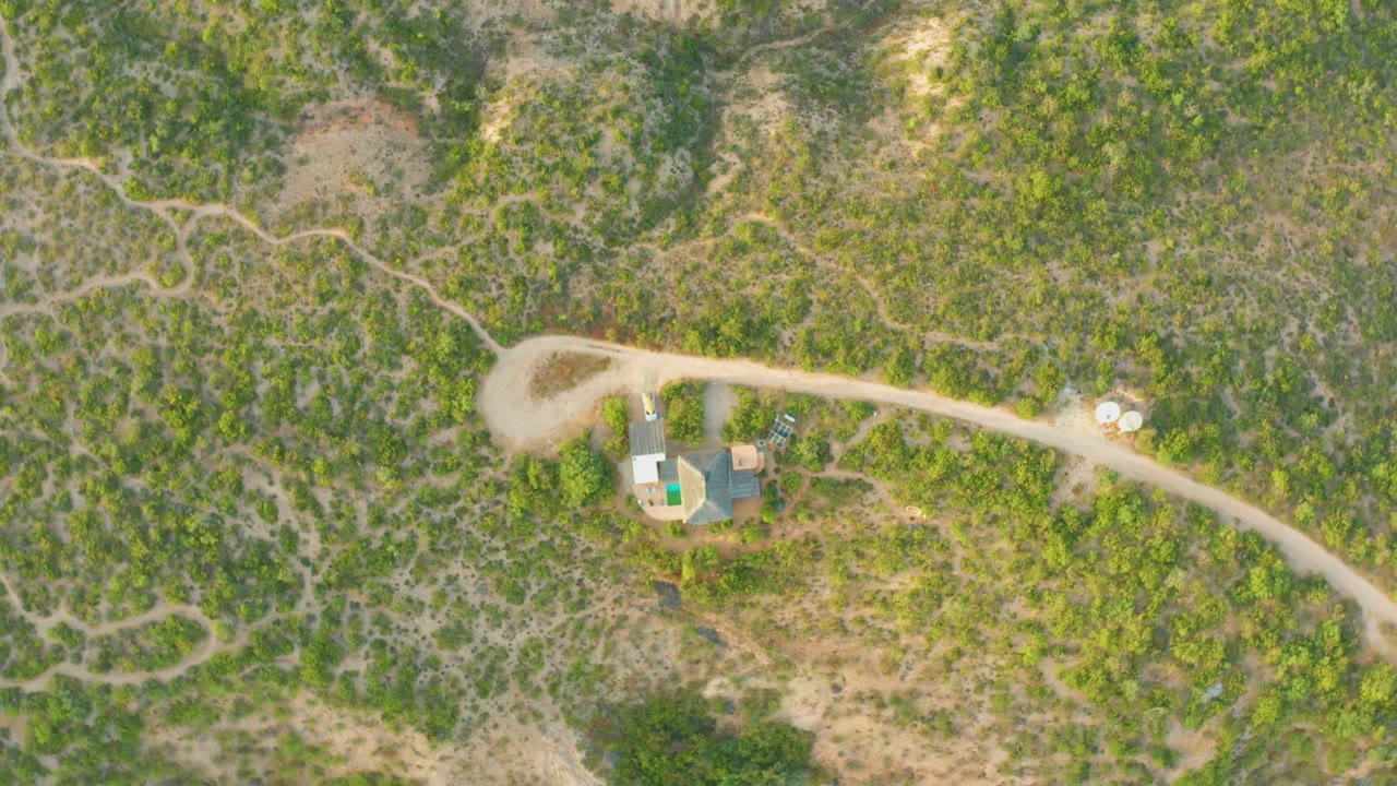 Drone-like overhead frame from Cabo Pulmo showing the dirt road curving past a cluster of small structures, with patchy vegetation and no visible people.