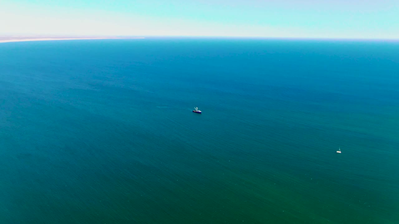 Aerial view over San Juanico, Baja California Sur, showing blue-green ocean, a small dark boat, a white buoy, a distant shoreline, and no visible people.
