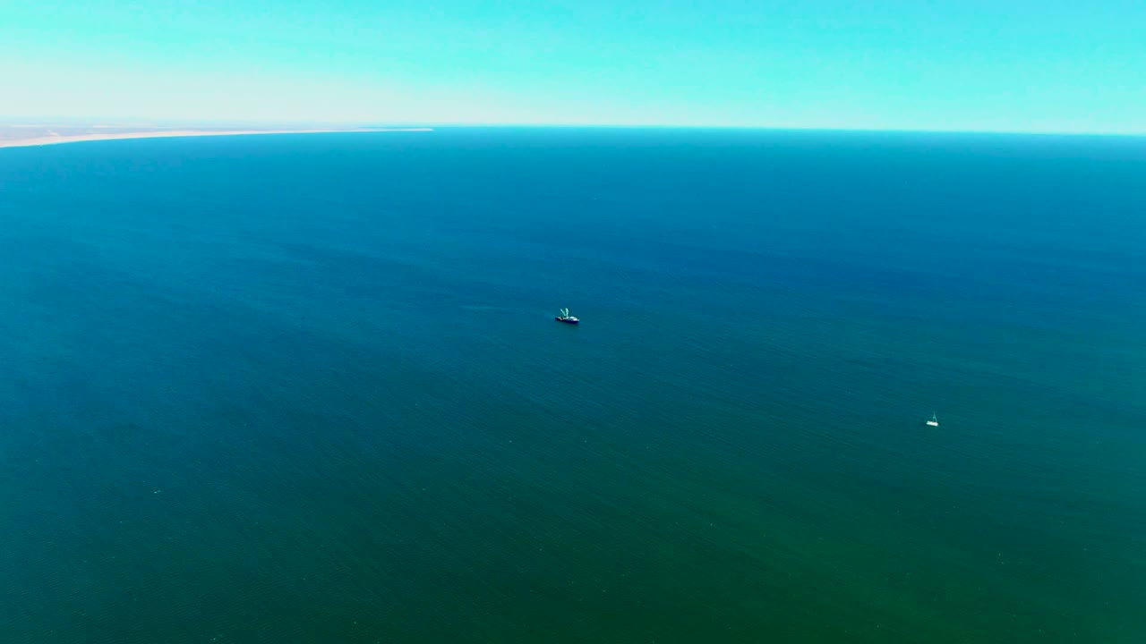 Aerial frame from San Juanico showing the small boat against darker blue-green water, a tiny white buoy to the right, distant coast, and no visible people.