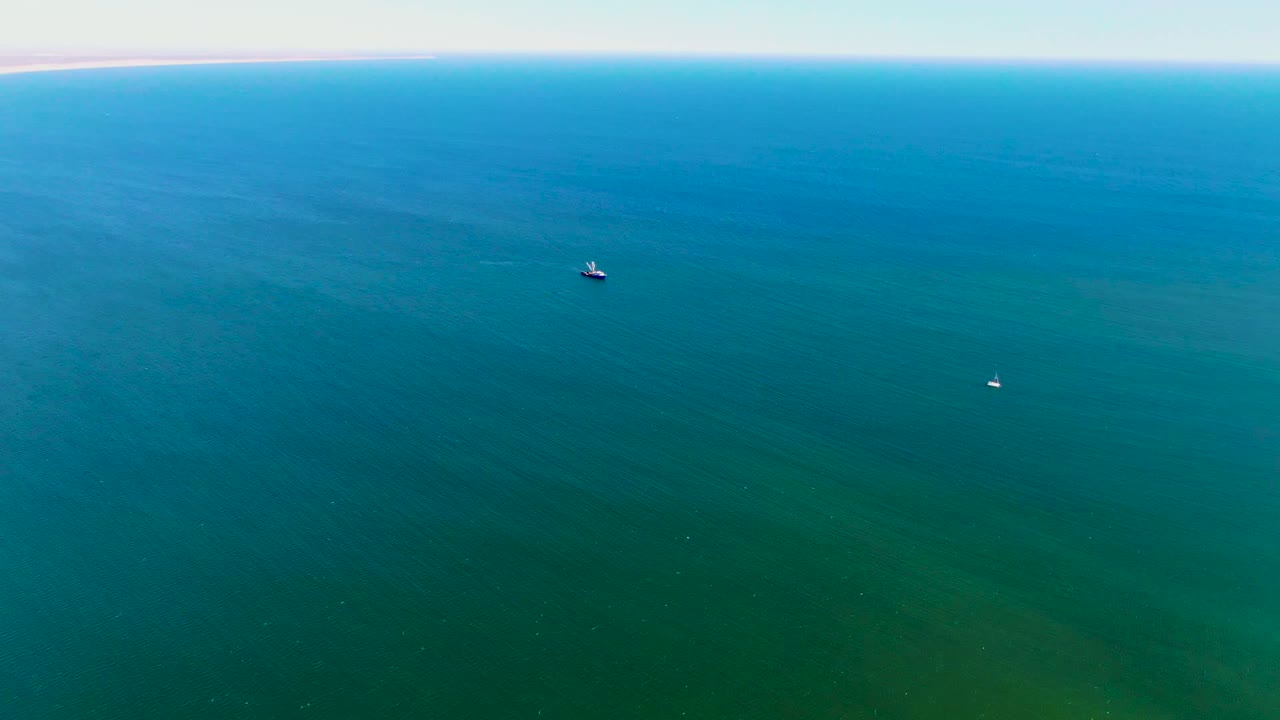 Wide aerial view over blue-green ocean near San Juanico, Baja California Sur, with a flat horizon, two small distant boats, and no visible people.