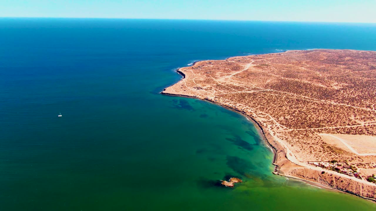 High aerial frame of San Juanico's sandy desert headland beside calm green and blue ocean, with sparse tracks on land and no visible people.