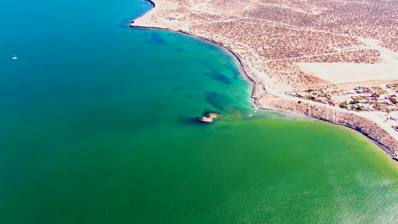 Aerial view over San Juanico, Baja California Sur, with sandy shoreline, small coastal buildings, green-blue water, and no visible people.
