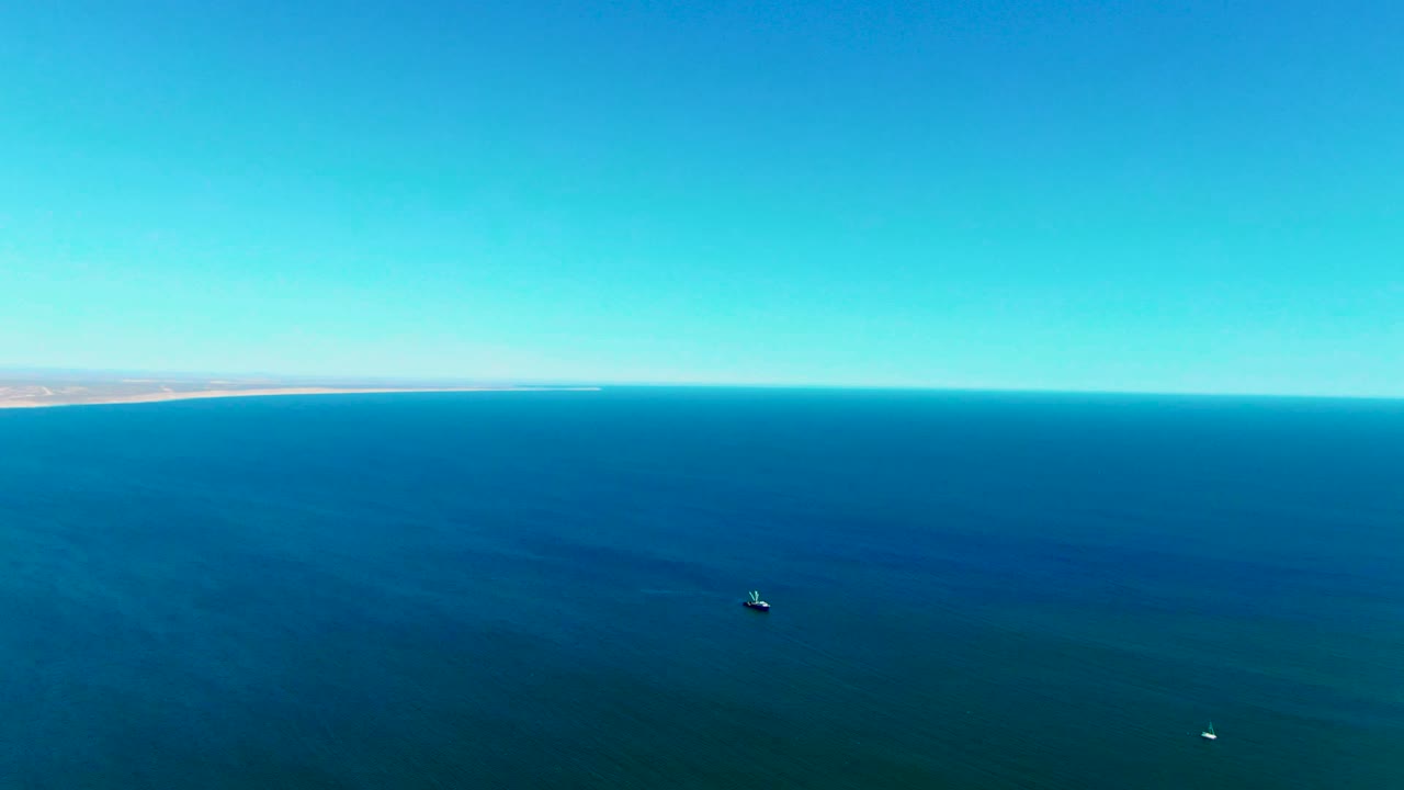 Aerial view over blue ocean near San Juanico, Baja California Sur, with a distant shoreline, two small boats on the water, clear sky, and no visible people.