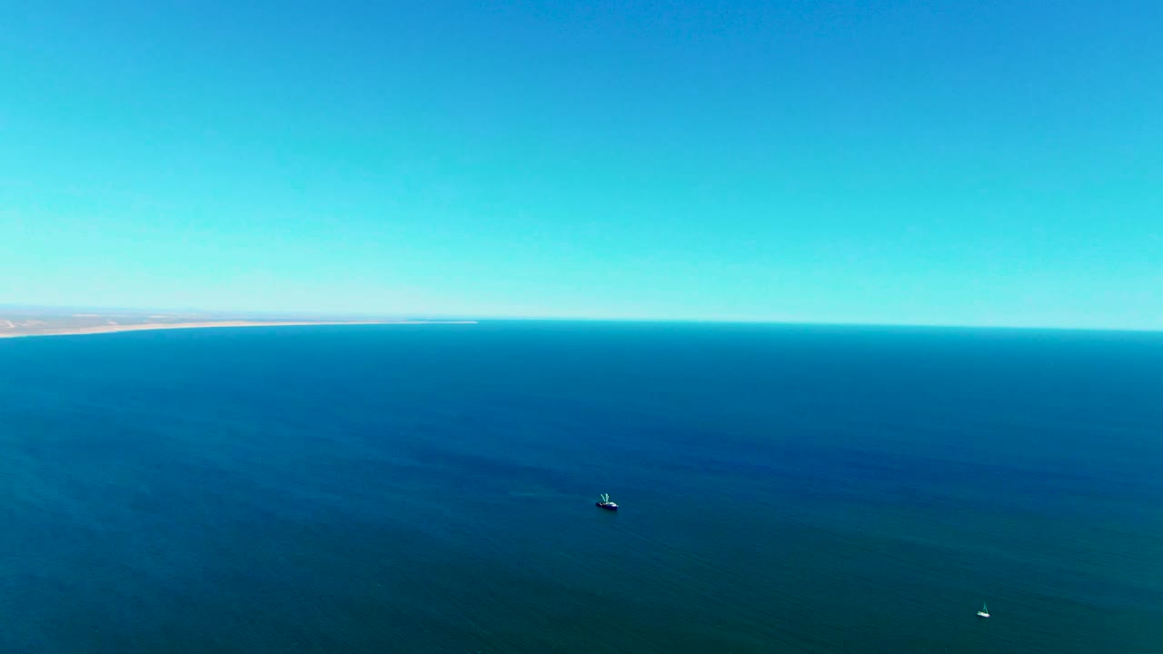 High view across the ocean near San Juanico, Baja California Sur, with the shoreline thin on the horizon, one small boat more central, a second small white boat lower in frame, and no visible people.