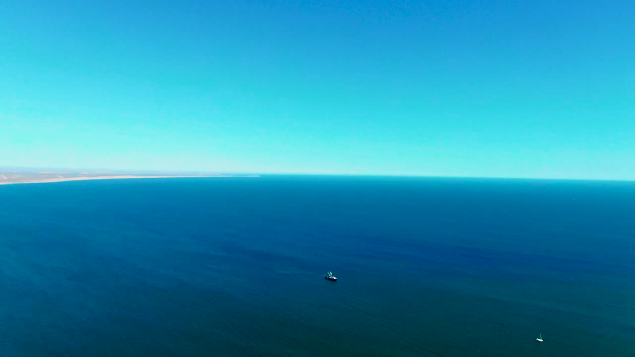 Aerial ocean frame near San Juanico, Baja California Sur, showing deep blue water, a level horizon, faint distant coast, two tiny boats spaced apart, and no visible people.