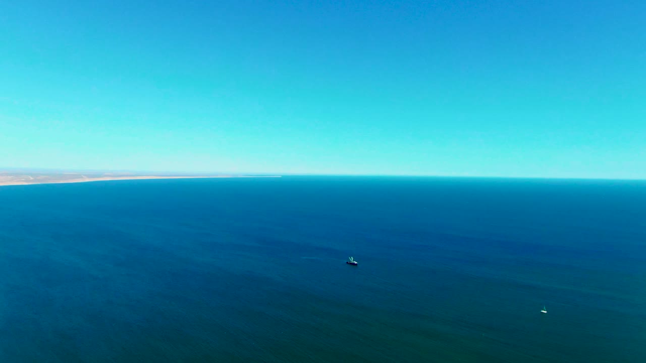 Wide aerial frame over the ocean at San Juanico, Baja California Sur, with the shoreline faint on the left horizon, a small dark vessel or buoy offshore, and no visible people.