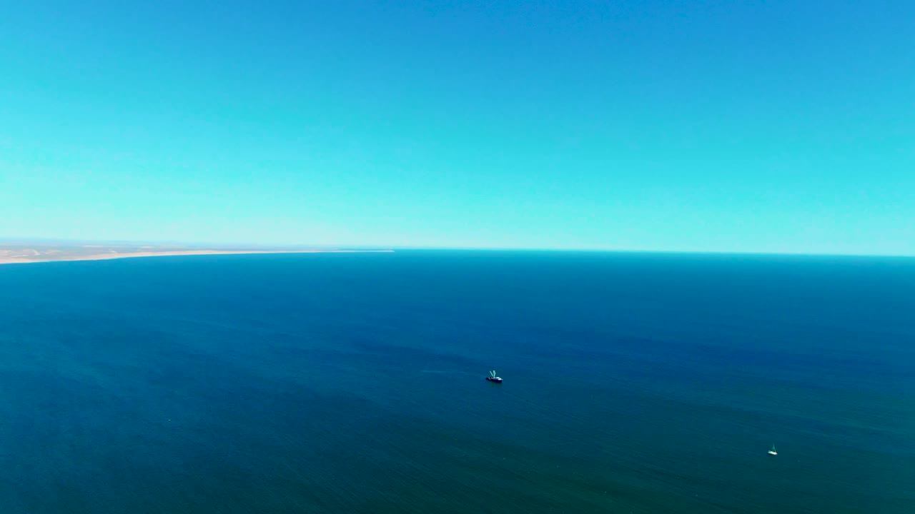 San Juanico aerial frame showing open blue water, the distant Baja California Sur shoreline at the left edge of the horizon, one small dark offshore object, and no visible people.