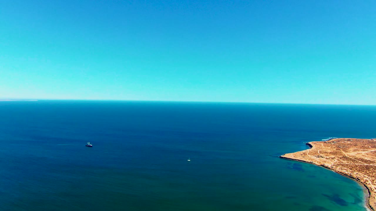 Aerial view over blue ocean near San Juanico, Baja California Sur, with a small boat on the left, a dry desert point entering from the right, clear sky, and no visible people.