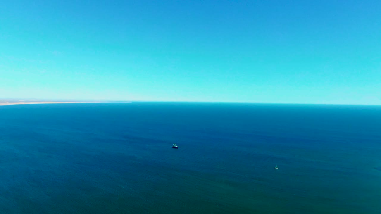 Aerial ocean view near San Juanico, Baja California Sur, with a small dark boat near center, a tiny white object on the water, distant land on the horizon, and no visible people.