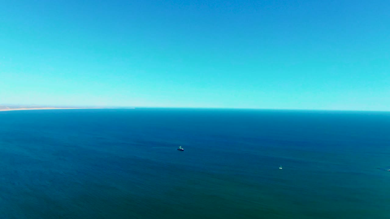 Wide aerial view over the ocean near San Juanico, Baja California Sur, with a clear blue sky, a level horizon, small boats on the water, and no visible people.