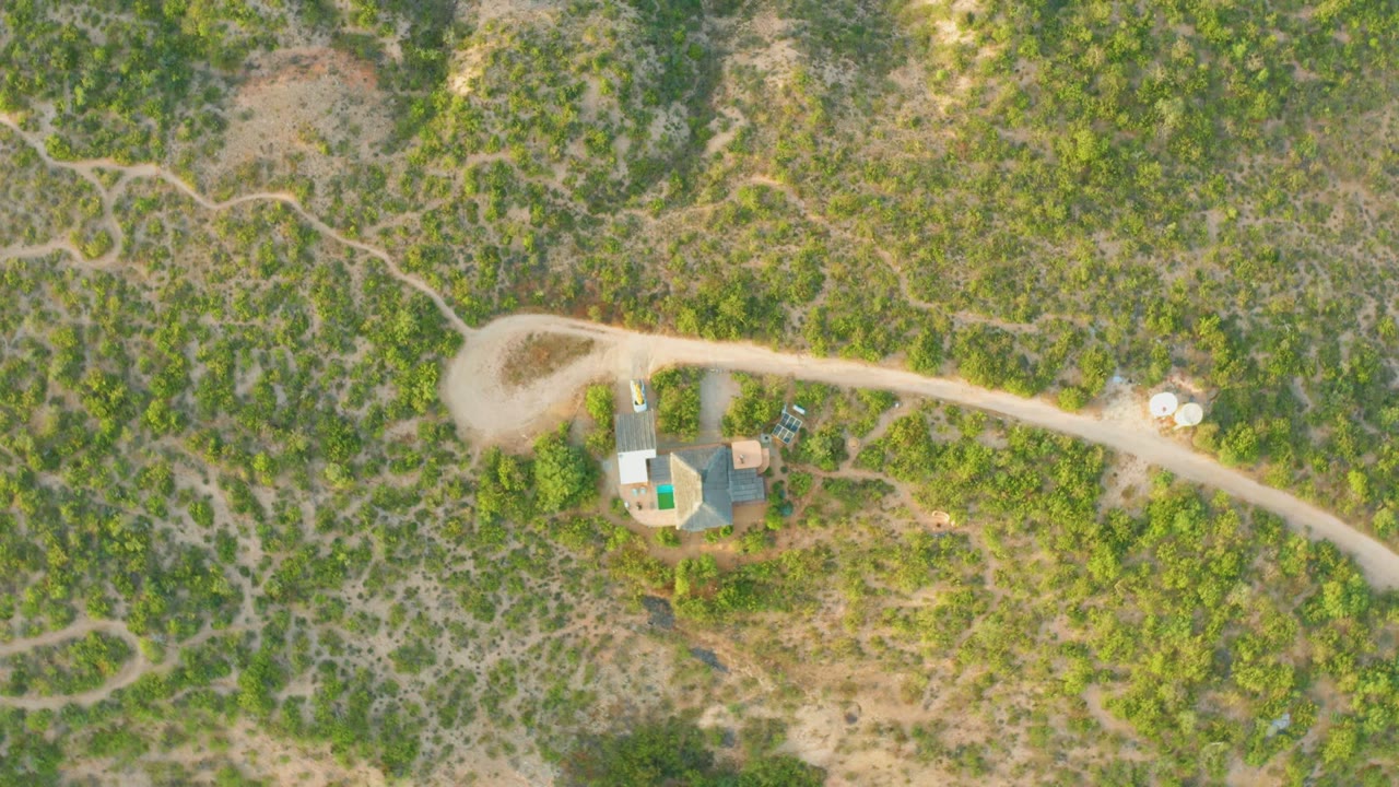 Overhead drone frame from Cabo Pulmo, Baja California Sur showing a house compound beside a pale dirt road, winding footpaths, scrub vegetation, and a tiny person near the buildings.