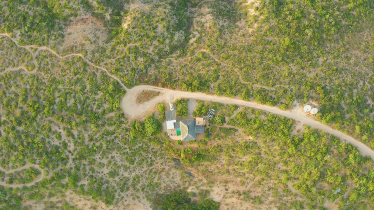 Overhead frame from Cabo Pulmo with the house cluster near the lower left, a pale road running across the frame, two light circular objects by the road, scrubby hills, and a tiny person near the structures.